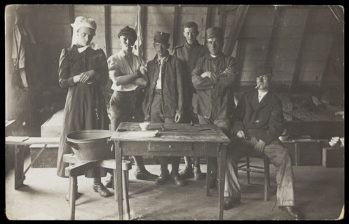 French prisoners of war, one in drag, gather round a small wooden table, [between 1910 and 1919?]. Creator: Unknown.