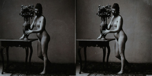 A young woman posing naked in a photographic studio, standing in front of a table on which..., c1900 Creator: Unknown.