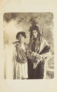 Two men in drag, one wearing a detailed feathered head garment, pose for a portrait, [1900-1909?]. Creator: Frank Wood.