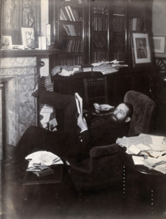 Arthur Schuster, lying on an armchair by a fireplace, with a photograph of Lord Kelvin..., c1901. Creator: Unknown.