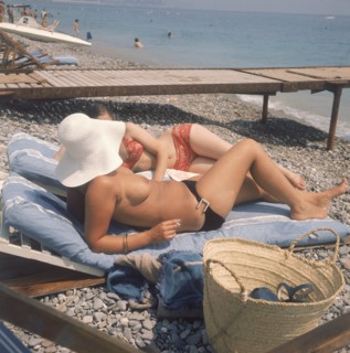 Dans les années 1970, deux femmes, dont l'une en monokini, bronzent sur une plage normande. Dans