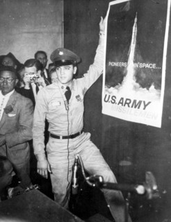Elvis PRESLEY as a soldier next to the poster 'Pioneers in Space' 1958