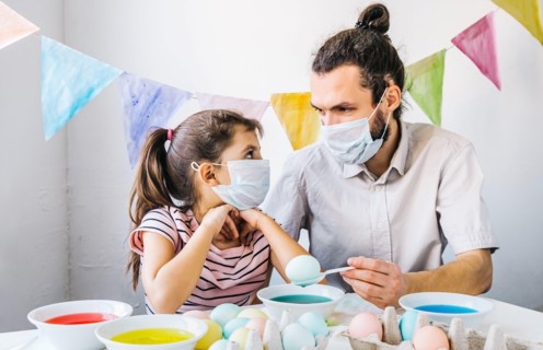 New normal Easter celebration lifestyle concept. Cute little girl and her dad wearing protective masks, preparing easter, dyed eggs at home during