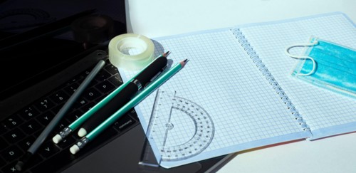 On the laptop keyboard lie a notebook pencils pens ball-point protractor adhesive tape and a disposable medical mask.
