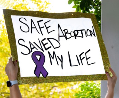 Sign with glitter edge reads Safe Abortion Saved My Live - blurred outdoor background at Reproductive Justice Rally