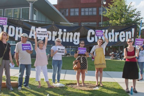 People hold pro women's health care and abortion signs in front of a large anti-abortion banner at a Moral Monday rally