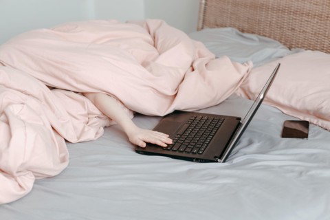 Bored funny child lying in bed under blanket and learning in virtual online school class. Hands typing on computer keyboard. Kid working on laptop