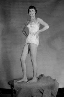 Young woman models a girdle while standing on a table, ca. 1955.