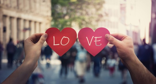 Two hands holding different paper heart symbols, unite them to create the word Love, on a crowded street. Woman and man togetherness concept. Valentin