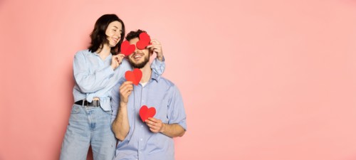 Flyer. Young and happy man and woman holding greeting cards shaped hearts isolated on pink trendy color background. Emotions, youth, love and