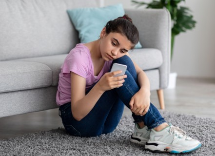Unhappy Indian teen girl with mobile phone feeling sad, suffering from cyber bullying online, sitting on floor at home