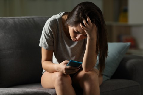 Sad teen being victim of cyber bullying online sitting on a couch in the living room at home