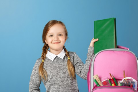 Closeup portrait of little girl schoolgirl on blue background. The child puts in a satchel book. Back to school. The concept of education.