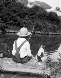 1920s 1930s FARM BOY WEARING STRAW HAT AND OVERALLS SITTING ON LOG WITH SPOTTED DOG FISHING IN POND