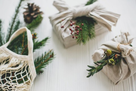 Stylish christmas gifts wrapped in linen fabric with green branch and reusable shopping bag with green spruce on rustic wooden background. Zero waste