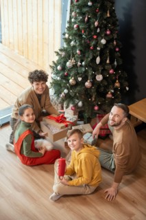 Portrait of positive beautiful Caucasian family sitting with presents on floor against Christmas tree at home