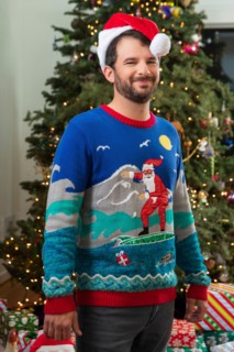 Portrait of a young man with a beard, wearing a Santa Hat and an ugly Christmas sweater standing in front of Christmas tree.