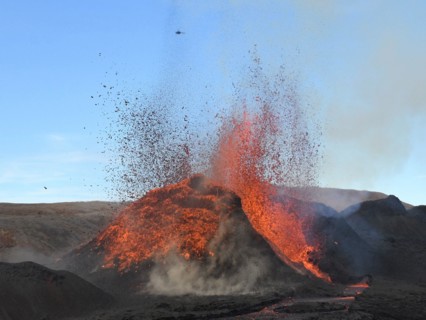Volcanic vent at Fagradalsfjall, Iceland, erupting incandescent orange and red lava. Black lava in the foreground and blue sky in the background.