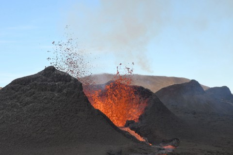 Volcanic vent at Fagradalsfjall, Iceland, erupting incandescent orange and red lava. Black cool lava in the foreground and blue sky in the background.