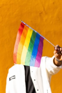 A young adult holds a multicolored rainbow flag in front of a bright yellow background