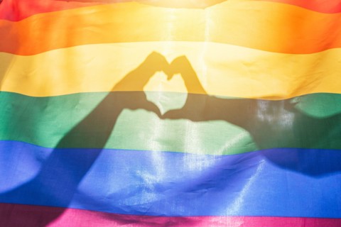 LGBTQ rainbow colors flag, a symbol for the homosexual community, two hands making a heart shape shining by sunlight. Gay flag for pride month and the