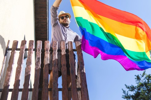 Man in hat holding lgbt rainbow flag in his hands while standing on the balcony.
