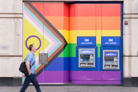 A man walks past two cash machines on the wall painted with rainbow colours during celebration of Pride Month England UK