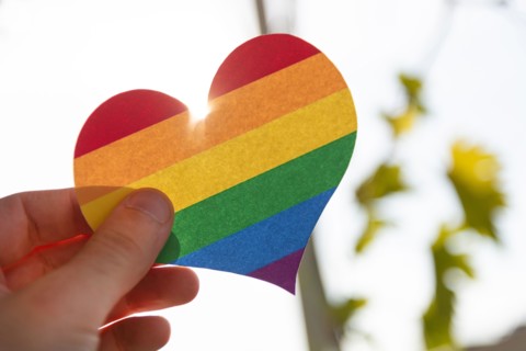 Hand holding a paper heart in LGBT rainbow colors with the sun rays shining around it, concept of LGBT, close-up