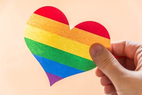 Hand holding a paper heart in rainbow colors or LGBT flags against a pink background, close-up