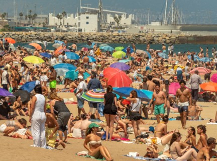 A bustling, crowded beach in Barcelona filled with people and colourful umbrellas, with the boats and breakwater of the port visible in the background.
