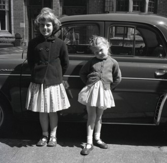1958, historical, two young girls, sisters, stand outside in a British street by their father's car, a French Renault Dauphine, a rear-engined 4-door economy car, a successor to the Renault 4CV.  Betw...