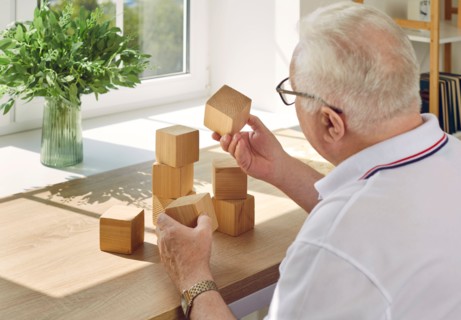 Elderly man collects wooden cubes at table opposite the window.