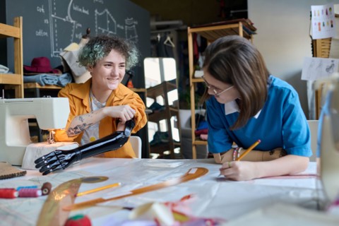 Portrait of two smiling teenage girls with prosthetics sewing clothing together in inclusive training class