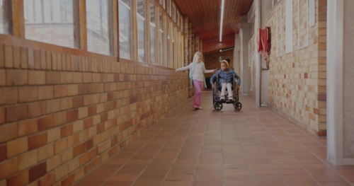 Two classmates rolling wheelchair and walking with backpack in school hall, with fire hose cabinet