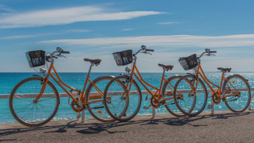 Three bright orange hire bicycles parked on the seaside promenade with an azur blue sea sparkling in the background