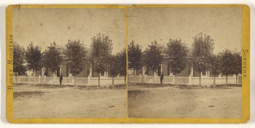 Rocky Mountain Scenery. Man in hat standing in front of a large tree-lined house, with white picket fence; American; about 1870