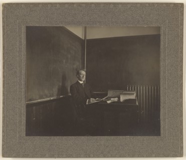 Portrait of a male teacher seated at his classroom desk; American; about 1900; Gelatin silver print