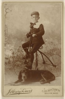 little boy wearing a cap, mounted on an old-style three-wheel bicycle, with a dog in the foreground; J. Henry Davis, American