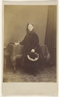 young girl in curls, standing, holding a hat, resting her other arm on a table with books on top; Joseph Brown, British, active