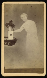 Female  spirit  standing next to a table with a photograph propped against a vase with flowers; William H. Mumler, American