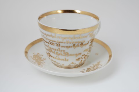 White cup and saucer fifty-year existence Inrigation for Women by Women with gold-colored rim and flower decoration, cup