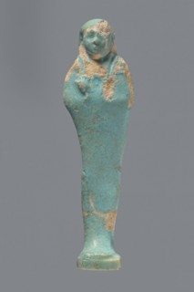 Ushabti. Mummy-shaped figurine standing on a base with a flat pillar at the back. Arms folded