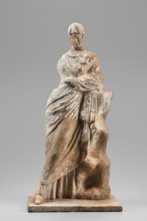 Female figurine. The figurine depicts a standing woman wrapped in a cloak, leaning her left elbow