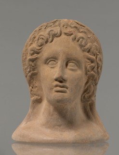 Votive female head, female head wearing a wreath, from under which lush locks of hair curl down the