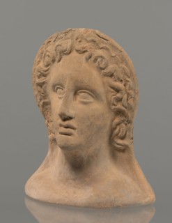 Votive female head, female head wearing a wreath, from under which lush locks of hair curl down the
