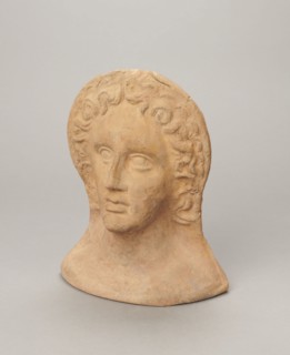 Votive female head, female head wearing a wreath, from under which lush locks of hair curl down the