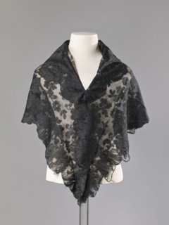Cape in the form of an elongated rhombus, made of black lace with floral motifs. The edge is