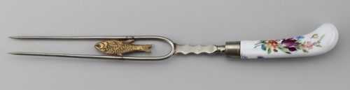 Two-pronged fork made of white metal with a gilded porcelain fish along the length between the