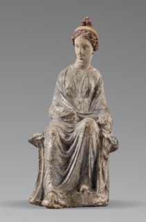 Figurine of a woman. A girl sitting in a chair is dressed in a soft white chiton over which a blue,