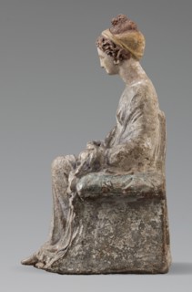 Figurine of a woman. A girl sitting in a chair is dressed in a soft white chiton over which a blue,